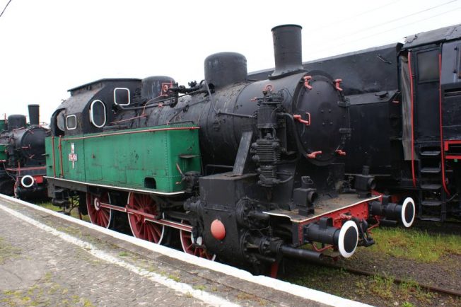 Stara lokomotywa parowa oznaczona numerem TKc100-1 stoi na torach kolejowych, za nią widoczne są inne wagony lub lokomotywy. Pojazd ma czarną kabinę i zielony tender, jest wyposażony w duże, czerwone koła oraz cylindryczny kocioł. Pomimo swojego wieku lokomotywa wygląda na zadbany eksponat z czasów historycznych kolei.