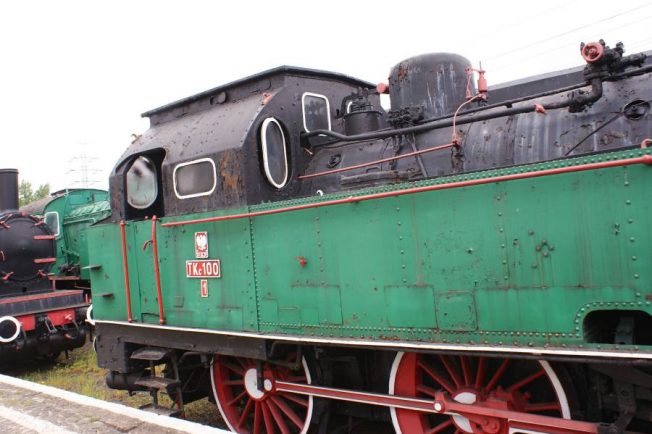 Czarno-zielona lokomotywa parowa z czerwonymi kołami stoi na torach; na bocznej stronie lokomotywy widnieje tabliczka z napisem 