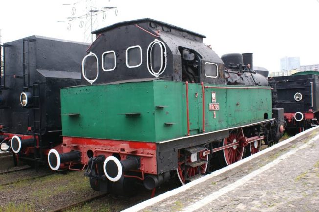 Czarno-zielona lokomotywa parowa z czerwonymi obramowaniami stoi na torach; jest wyposażona w komorę ogniową, kocioł, kominy i koła parowe. Tuż obok lokomotywy znajduje się inna lokomotywa w podobnym kolorze. Obie maszyny są umiejscowione na otwartej przestrzeni z twardą nawierzchnią przypominającą peron lub plac manewrowy.