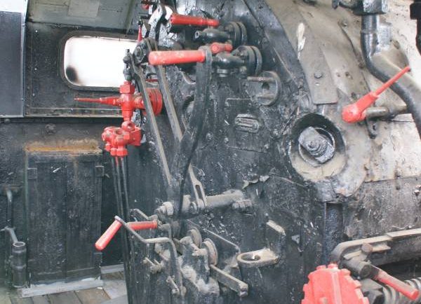 Kabina maszynisty lokomotywy parowej z wyróżniającymi się czerwonymi elementami, takimi jak dźwignie i pokrętła. W środku widoczne są liczne urządzenia i wskaźniki używane do sterowania parowozem. Przestrzeń jest funkcjonalna i zawiera niezbędne wyposażenie do obsługi maszyny.