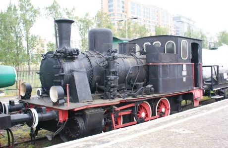 The black steam locomotive with red-rimmed wheels and fittings stands on the tracks. It has a classic design with a wide cab enclosure and a tall chimney. In the background, other rail vehicles and urban buildings are visible.