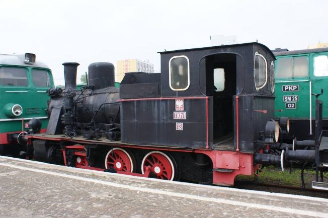 Czarno-czerwona lokomotywa parowa z tendrem stoi na torach obok innych pojazdów szynowych. Ma białe obramowania wokół okien i na przedniej części lokomotywy, gdzie widnieje także numer 