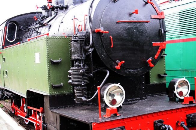 Część zielono-czerwonej lokomotywy parowej jest widoczna, z dużym okrągłym czarnym kotłem i reflektorami. Na boku kotła znajdują się czerwone obramowania i zawory. Również widać elementy mechanizmu napędowego i koła z szerokimi czerwono czarnymi obręczami.