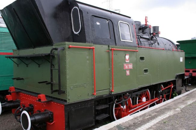 Zielono-czerwona lokomotywa parowa z dużym czarnym kominem stoi na torach. Maszyna ma na boku napis 