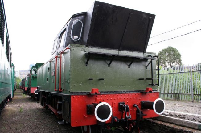 Zielono-czarna lokomotywa parowa z czerwonymi obramowaniami stoi na torach; widoczne są koła, wejście do kabiny oraz kominek. Po lewej stronie lokomotywy znajduje się ciąg innych wagonów zielonego koloru. Tło stanowi nieco zasnuta chmurami szara niebo oraz elementy infrastruktury kolejowej.