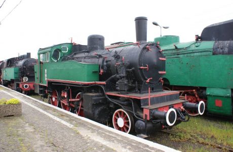 The black steam locomotive with red trim is standing on a side railway track next to other green locomotives. The machine has numerous pipes, valves, and brass elements that are characteristic of old steam locomotive designs. In front of the locomotive, there is a platform with a white line, suggesting a place for visitors or for staff to work.