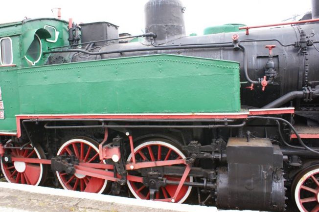 Zielona lokomotywa parowa z czerwonymi obręczami kół eksponowana jest na zewnątrz. Maszyna posiada czarny kocioł, duży komin oraz system rur i zbiorników. Na bokach widnieje czarny podwozie z metalowymi elementami, które są częścią układu napędowego.