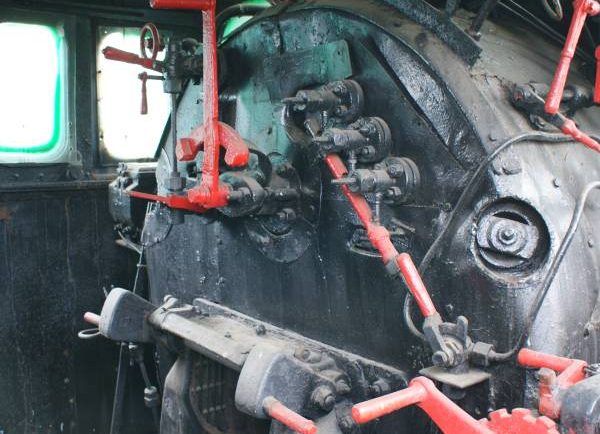 Widzimy wnętrze starej lokomotywy parowej z widocznymi elementami sterowania i wskaźnikami. Ręczne uchwyty i regulatory są pomalowane na czerwono, a kotły i rury w tle mają odcień czerni i są zrobione z metalu. Ściany kabiny są drewniane, a przez okno wpada naturalne światło.