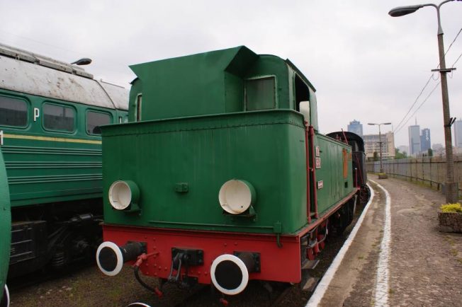 Lokomotywa parowa o zielono-czerwonym malowaniu stoi na torach kolejowych. Posiada okrągłe, białe reflektory i duży czarny kocioł, typowy dla dawnych parowozów. W tle widoczne są inne pojazdy kolejowe oraz miejski krajobraz, w tym wysokie budynki.
