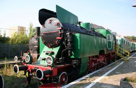 The green and black steam locomotive stands on the tracks; it has red trim and white wheels. The sides show elements of the driving mechanism and large round lanterns. The locomotive is well-preserved, and in the background, other rail vehicles can be seen.