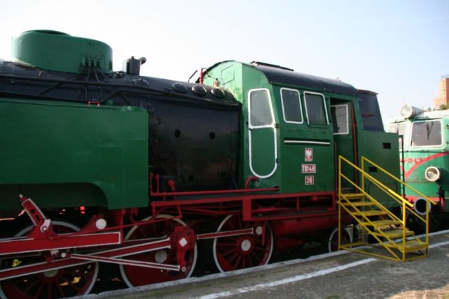 Zielona lokomotywa parowa z dużymi, czerwonymi kołami stoi na torach obok platformy z żółtymi schodami. Na maszynie widoczne są białe oznaczenia 