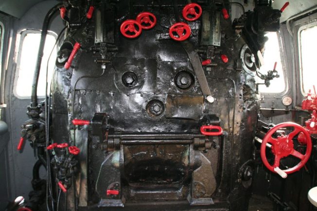 Widok wnętrza kabiny lokomotywy parowej z różnymi urządzeniami kontrolnymi. Dominują kolory ciemnoszare, czarne oraz akcenty czerwone na pokrętłach i zaworach. Ściany i elementy są metaliczne z widoczną patyną użytkowania.