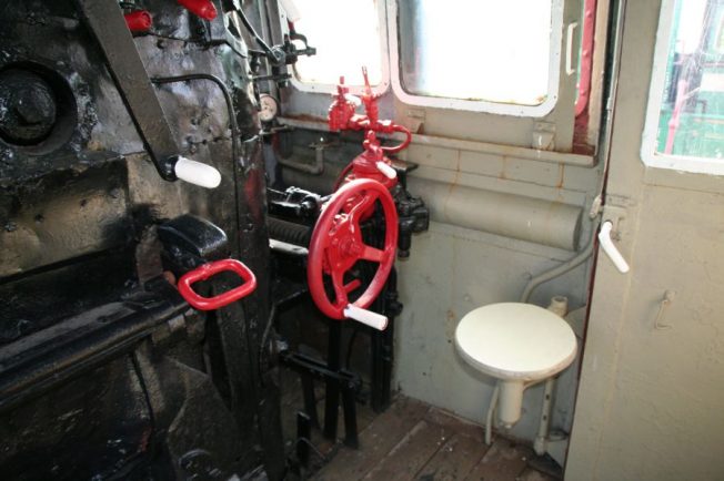 Kabina lokomotywy pokazuje pulpit sterowniczy z licznymi wskaźnikami i dźwigniami pomalowanymi na czerwono. Przestrzeń jest ograniczona, z widocznym, umieszczonym centralnie, okrągłym taboretem. Na ścianach kabiny zauważalne są elementy metalowe i okno.