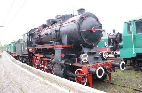 The black steam locomotive with red elements stands on the railroad tracks, surrounded by dry grass and several other railway vehicles. The large wheels of the locomotive, pipes, and complex mechanical structure characteristic of steam locomotives are visible. Three round headlights are mounted on the front of the locomotive, and white markings are found on the side walls.