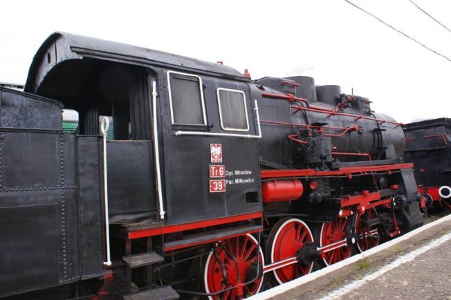 Czarno-czerwona lokomotywa parowa z bocznymi czerwonymi kołami i czarnym kotłem stoi na torach. Na boku kabiny maszynisty wisi tabliczka z napisem 