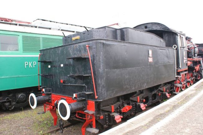 Czarny parowóz stoi na torach kolejowych obok zielonego wagonu. Posiada czerwone obramowania na kołach oraz białe linie podkreślające poszczególne elementy konstrukcji. Konstrukcja parowozu z dużym tendrem wskazuje na starą technologię parową stosowaną w lokomotywach.