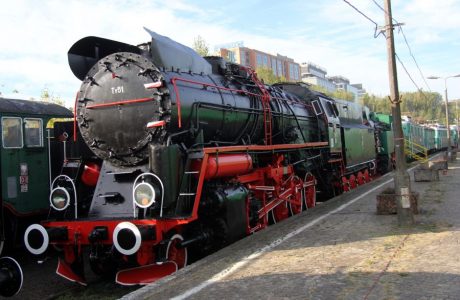 The black and red steam locomotive is standing on the tracks next to the platform, surrounded by other railway vehicles. It has a round, shiny front buffer, numerous pipes and fittings, and on the side, a large letter "T" and the numbers "51-228" are visible. It is characterized by a sturdy construction and a classic steam locomotive appearance with a large, round chimney and horizontal cylinders.