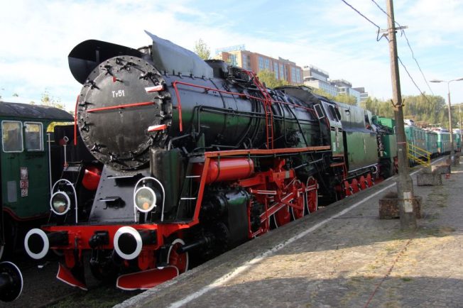 Czarno-czerwona lokomotywa parowa stoi na szynach obok peronu, otoczona innymi pojazdami kolejowymi. Posiada okrągły, lśniący przedni bufor, liczne rury i okucia, a na boku widać wielka literę 
