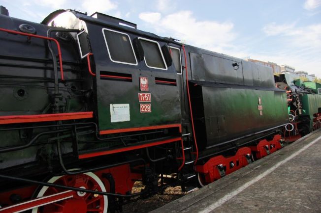 Czarna, zielona z czerwonymi akcentami lokomotywa parowa stoi na torach. Maszyna posiada tabliczkę z napisem 