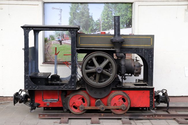 Czarno-czerwona spalinowa lokomotywa o niewielkich rozmiarach stoi na torach. Maszyna posiada jedną kabinę z oknami i proste, metalowe pręty ochronne po bokach. Lokomotywa wyposażona jest w duże, centralnie umieszczone koło napędowe i mniejsze koła wspierające.