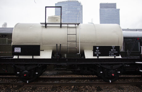 Dwuosiowy wagon cysterna stoi na torach kolejowych, wyposażony jest w nitowany zbiornik o cylindrycznym kształcie. Po bokach wagonu znajdują się identyfikacyjne tabliczki z napisami. Tło obrazu wypełniają kontury budynków miejskich z lekko zachmurzonym niebem.