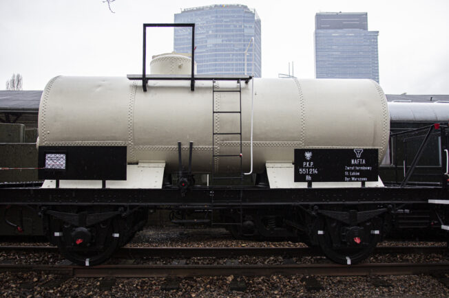 Dwuosiowy wagon cysterna stoi na torach kolejowych, wyposażony jest w nitowany zbiornik o cylindrycznym kształcie. Po bokach wagonu znajdują się identyfikacyjne tabliczki z napisami. Tło obrazu wypełniają kontury budynków miejskich z lekko zachmurzonym niebem.