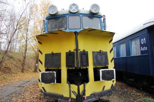 Żółto-czarny pług odśnieżny stoi na torach obok niebieskiego wagonu kolejowego. Konstrukcja ma dwa wirniki umieszczone centralnie, z przodu, oraz dodatkowe oświetlenie umieszczone wyżej. Po bokach znajdują się składane stopnie umożliwiające dostęp do kabiny.