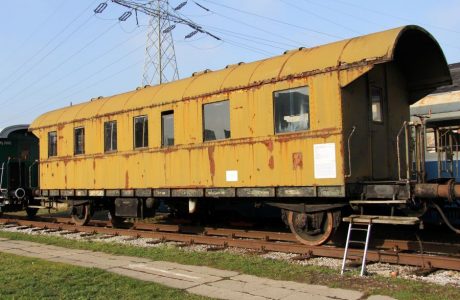 Stary, żółty wagon kolejowy stoi na bocznych torach. Wagon posiada dach w kształcie łuku i jest wyposażony w kilka okien oraz drzwi. Pojazd wygląda na nieeksploatowany od jakiegoś czasu, brak na nim oznakowania użytkowego.