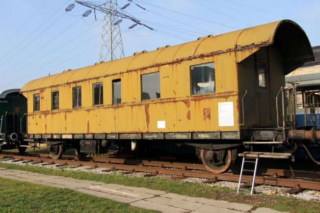 Stary, żółty wagon kolejowy stoi na bocznych torach. Wagon posiada dach w kształcie łuku i jest wyposażony w kilka okien oraz drzwi. Pojazd wygląda na nieeksploatowany od jakiegoś czasu, brak na nim oznakowania użytkowego.