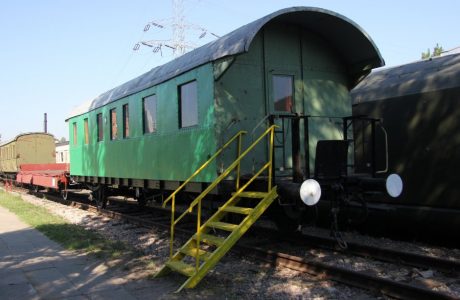 Widoczny jest zielony wagon pasażerski stojący na torach kolejowych, wyposażony w metalowe schody przy wejściu. Wagon posiada okrągłe okna oraz charakterystyczne, kształtne zderzaki. Po lewej stronie wagonu widać fragment innego pojazdu kolejowego o czerwonym kolorze.