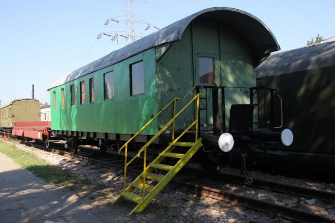 Widoczny jest zielony wagon pasażerski stojący na torach kolejowych, wyposażony w metalowe schody przy wejściu. Wagon posiada okrągłe okna oraz charakterystyczne, kształtne zderzaki. Po lewej stronie wagonu widać fragment innego pojazdu kolejowego o czerwonym kolorze.