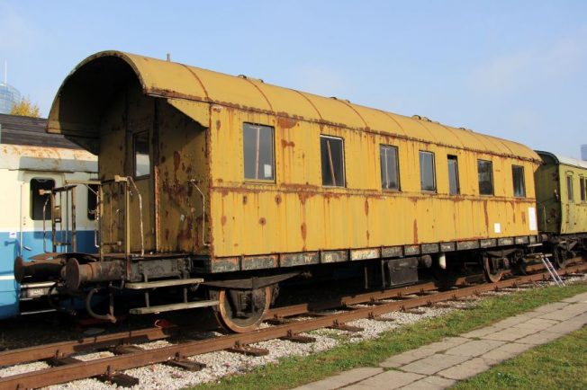 Żółty wagon pasażerski stoi na torach, ma wiele okien i drzwi po obu stronach. Konstrukcja metalowa z widocznymi elementami jak drabinki i haki. Wagon otacza spokojny teren z trawą i innymi pojazdami kolejowymi w tle.