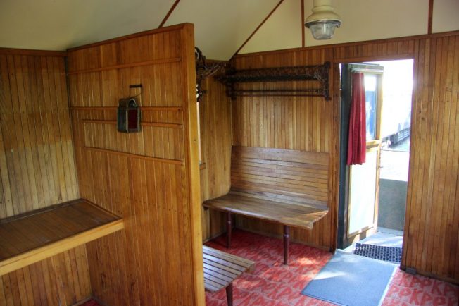 Wagon pasażerski typu BCi-29 ukazuje wnętrze z drewnianymi ścianami i ławkami przeznaczonymi dla pasażerów. Na podłodze widoczny jest czerwony dywan, a wnętrze oświetla wisząca lampa. Przy drzwiach znajduje się metalowa poręcz oraz stopnie ułatwiające wejście do wagonu.