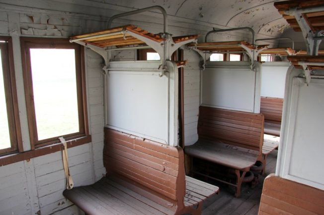 Wnętrze przedstawia drewniane siedzenia rozmieszczone naprzeciwko siebie w wagonie kolejowym, z miejscami dla pasażerów podzielonymi niskimi drewnianymi ściankami. Nad siedzeniami znajdują się otwarte półki na bagaż, a okna są duże i zapewniają dobre oświetlenie kompartmentu. Ściany i sufit mają jasne odcienie, co tworzy przytulną atmosferę.