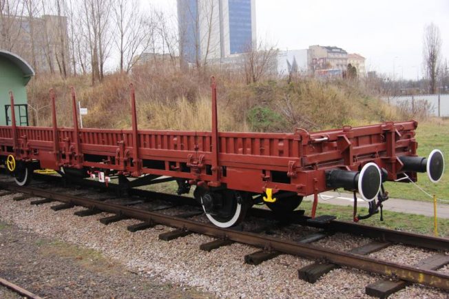 Czerwony wagon platforma stoi na torach kolejowych; posiada dwie osie i boczne ściany mogące służyć za barierki ochronne. Wagon wyposażony jest w metalowe elementy takie jak hak sprzęgający i ręczne kółka hamulcowe. Na tle wagonu widać trawnik i fragmenty miejskiej architektury, w tym budynek.