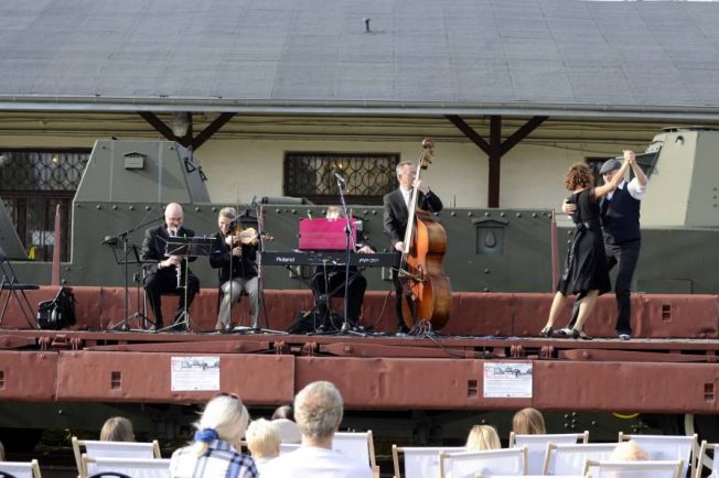 Na długim, metalowym wagonie platformowym stoi pięcioosobowy zespół muzyczny i wykonuje koncert na instrumentach takich jak: skrzypce, klarnet, kontrabas i puzon. W widowni siedzi grupa osób skierowana w stronę muzyków, a w tle widać zielone drzewa i fragment innych wagonów kolejowych. Koncert odbywa się na zewnątrz w słoneczny dzień, a widzowie wydają się zrelaksowani i skupieni na występie.