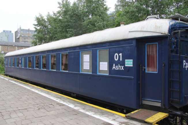 Niebieski wagon kolejowy stoi na peronie, oznaczony numerem 01 i oznaczeniem Ashx. Posiada białe napisy oraz czarne podwozie, a jego dach jest pokryty lekkim nalotem. Okna wagonu są przyciemniane, a przez otwarte można zauważyć wnętrze.