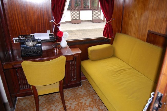 Wnętrze przedstawia elegancko urządzony wagon salonowy z żółtą sofą, drewnianym stołem i krzesłem. Na stole stoi maszyna do pisania i wazon z czerwonymi kwiatami, a okno zasłaniają czerwone zasłony. Podłoga pokryta jest wzorzystą wykładziną, a ściany wyłożone są ciemnym drewnem.