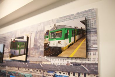 Na fotografii zawieszonej na ścianie widoczny jest zielony pociąg stojący na peronie stacji kolejowej. Obrazek ten jest jednym z wielu eksponatów prezentujących rozwój mazowieckich kolei. W tle za szklaną ochroną zawieszone są inne zdjęcia związane z tematyką kolejnictwa.