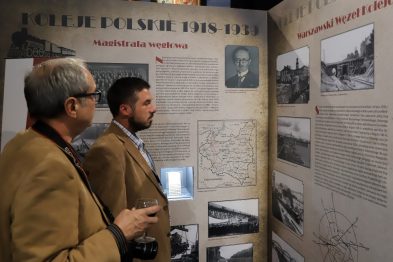 Dwóch mężczyzn ogląda ekspozycję poświęconą historii polskich kolei z okresu 1918-1939. W tle widoczne są historyczne fotografie, mapy i teksty opisujące rozwój kolejnictwa. Oświetlone witryny i ściany pokryte materiałem informacyjnym tworzą edukacyjną przestrzeń.