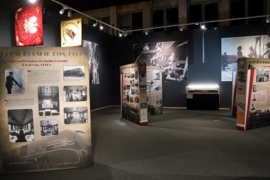 Eksponaty historyczne prezentujące rozwój polskich kolei w okresie międzywojennym są wystawione w muzeum; widać fotografie, dokumenty i plakaty na ścianach oraz na wolnostojących panelach. Sale muzealne są oświetlone, a wystrój wnętrza wydaje się być dopracowany, co podkreśla ważność prezentowanych zbiorów. Na jednym z paneli widoczny jest wizerunek orła – symbolu Polski.