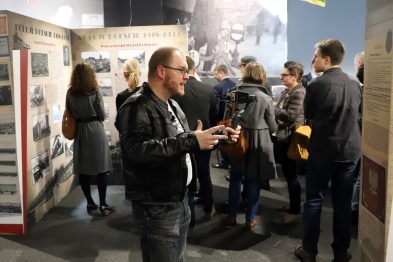 Grupa osób ogląda wystawę poświęconą kolei, stojąc przed planszami z fotografiami i informacjami. Mężczyzna w okularach na pierwszym planie trzyma telefon komórkowy, być może dokumentując ekspozycję. W tle widać różnorodne eksponaty, a także inne osoby zajęte zwiedzaniem.