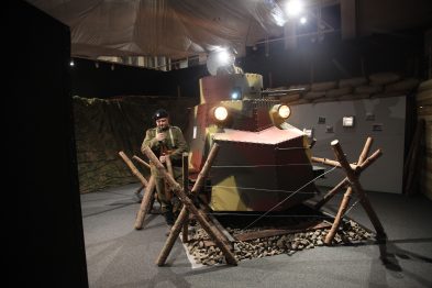 W pomieszczeniu oświetlonym reflektorami stoi rekonstrukcja lokomotywy pancernej z czasów historycznych, umiejscowiona na sztucznych torach kolejowych. Przed lokomotywą umieszczone są drewniane przeszkody w postaci ściętych pni, a całość jest otoczona kamuflażową siatką. Osoba w mundurze wojskowym siedzi obok lokomotywy, co nadaje ekspozycji realistyczny charakter.