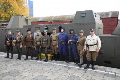 Grupa osób ubrana w historyczne mundury wojskowe stoi przed zielonym wagonem pociągu pancernego. Osoby te mają różne stroje, odzwierciedlające różne epoki i role wojskowe, niektórzy trzymają hełmy lub inne akcesoria. W tle widoczne są drzewa z liśćmi w jesiennych kolorach oraz fragmenty innych pojazdów.