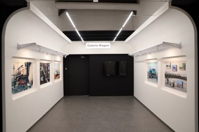 Widoczne jest wnętrze galerii ze zdjęciami zawieszonymi na białych ścianach, czarne ramy kontrastują z jasnym otoczeniem. W centralnym punkcie galerii znajduje się ciemne przejście prowadzące do kolejnych części ekspozycji. Oświetlenie galerii jest punktowe i skierowane na fotografie, co podkreśla ich detale i kolory.