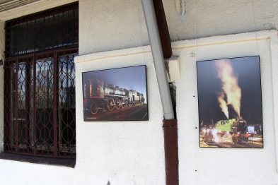 Dwa duże zdjęcia parowozów są zawieszone na zewnętrznej ścianie budynku obok okiennych krat. Na jednym fotografii widać maszynę w ruchu nocą, emitującą dym i parę, podświetloną z dołu. Druga fotografia ukazuje parowóz na tle czerwonego nieba, z wyraźnymi konturami i wydobywającym się dymem.