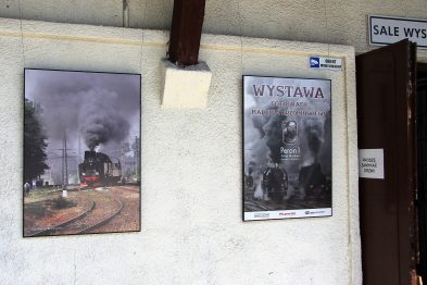 Na ścianie wisi kilka dużych fotografii przedstawiających parowozy w ruchu, emitujące obłoki pary i dymu. W centrum uwagi jest plakat informacyjny o wystawie fotografii Marcina Brzomińskiego. W tle widać wejście do budynku z oznaczeniem „sala wystawowa”.