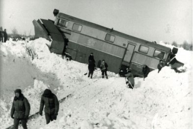 Pociąg osobowy wykoleił się i leży na boku na zboczu przykrytym śniegiem. Osoby w ciemnych kurtkach chodzą w pobliżu i przyglądają się sytuacji. Śnieg pokrywa większość terenu wokół torów, a niebo jest zachmurzone.