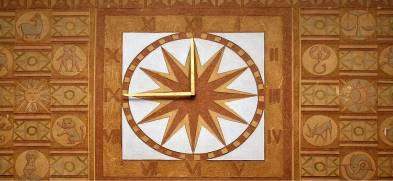 Zegar w kształcie koła z wachlarzowym wzorem znajduje się na pośrodku dekoracyjnej, geometrycznej ściany. Wyraźne złoto-brązowe kolory zdobią tło, które składa się z kwadratowych płytek z rozmaitymi wzorami. Wskazówki zegara są schludnie ustawione, wskazują konkretną godzinę.