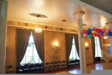 Wewnątrz eleganckiego pomieszczenia z wysokimi sufitami zdobione są złotymi ornamentami oraz ozdobnymi listwami przy stropie. Duże okna są przykryte ciężkimi zasłonami, a ściany zdobią dekoracyjne balony. Widoczne są krzesła ustawione wzdłuż ściany oraz klatka schodowa prowadząca na piętro.